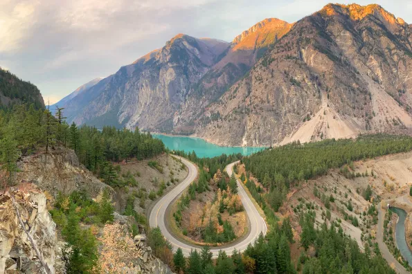 An aerial view of a road winding through the mountains with a bright greenish blue lake in the distance
